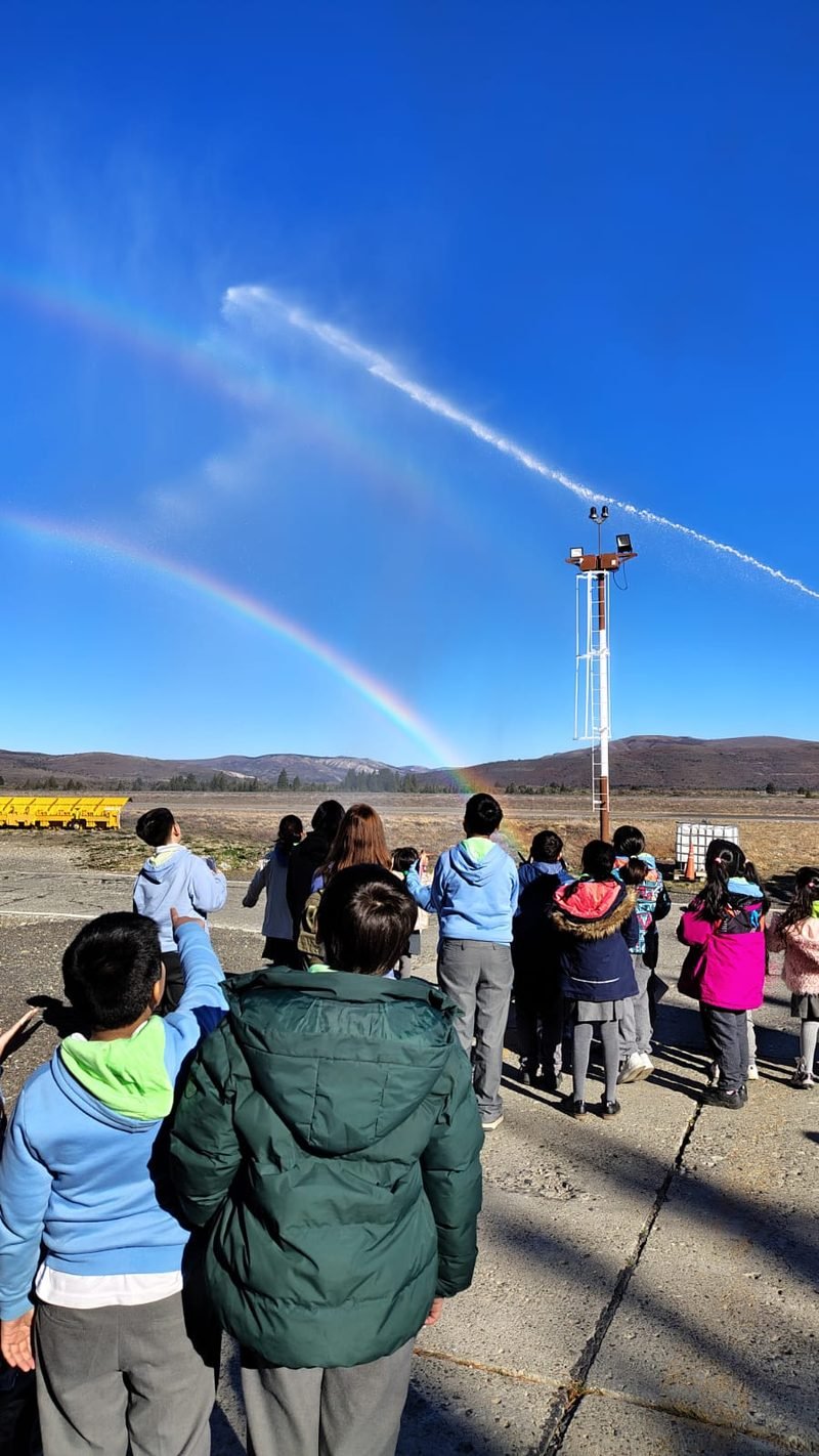 Estudiantes de espaldas miran un arcoíris que forma un gran chorro de agua en un campo abierto, bajo un cielo azul
