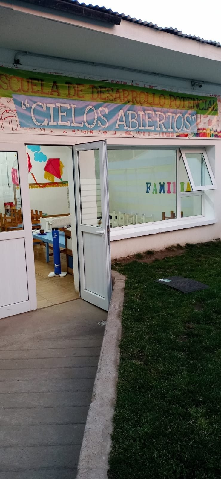 Entrada del edificio de la escuela con el cartel «Cielos Abiertos» y la puerta abierta junto al césped