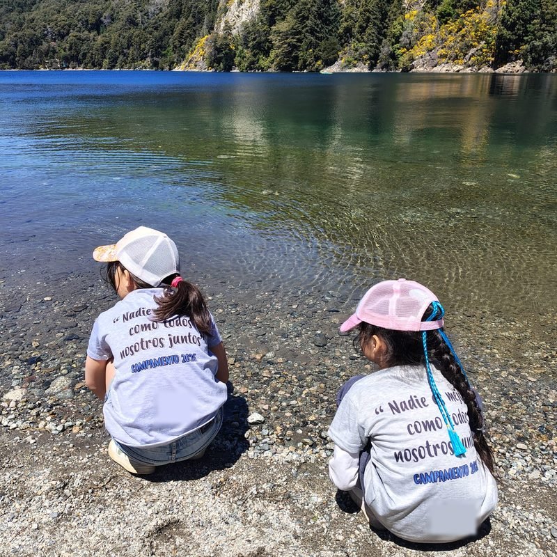 Dos alumnas de Cielos Abiertos de espaldas, con gorras y remeras del campamento, agachadas en la orilla de piedras de un lago de agua transparente rodeado de bosque