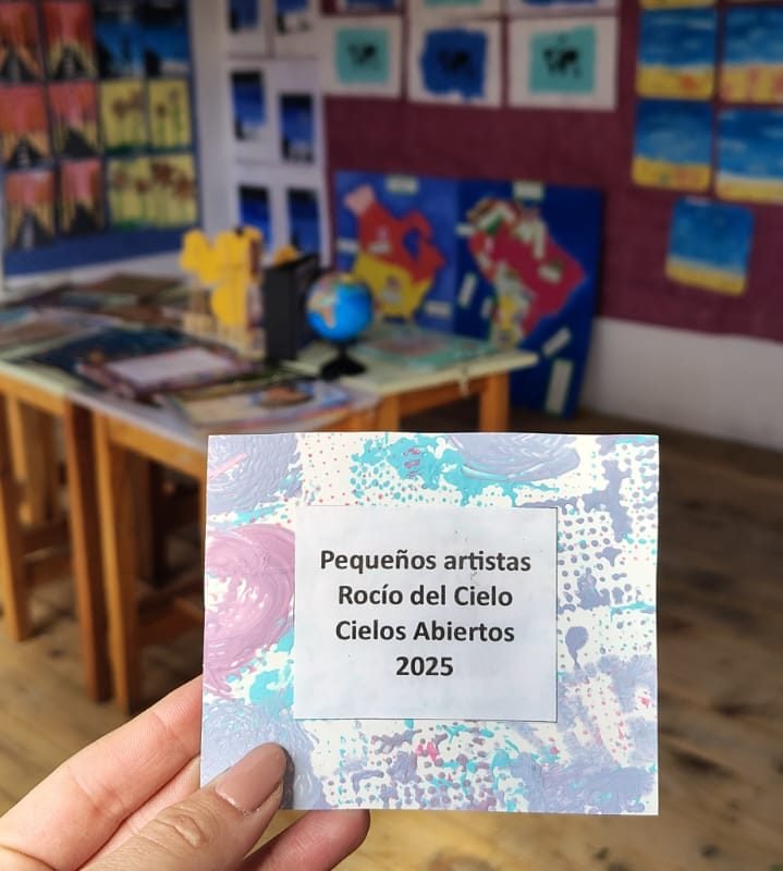 Una mano sostiene la tarjeta «Pequeños artistas. Rocío del Cielo, Cielos Abiertos 2025» frente a los trabajos de la muestra colgados en el aula