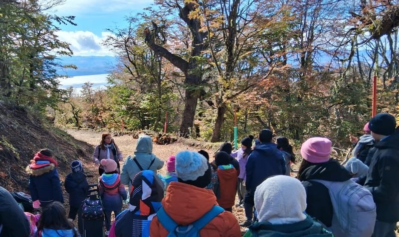 Grupo de estudiantes abrigados y con mochilas, de espaldas, en un sendero de bosque con árboles de otoño y el lago al fondo
