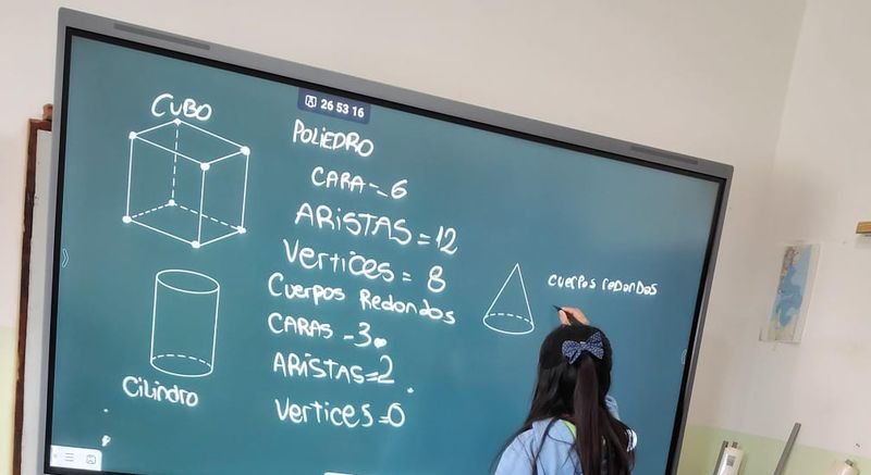 Una alumna de espaldas escribe en la pantalla inteligente las caras, aristas y vértices del cubo y del cilindro